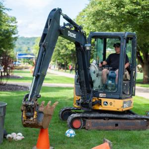 A student operates a small John Deere excavator, attempting to scoop a soccer ball off the top of a traffic cone.