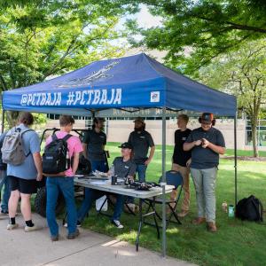 Students wearing golf shirts that say "PCT Baja" sit and stand behind a table under a blue canopy that also says "PCT Baja." Next to the table is a single-seat offroad vehicle the club builds and races each year. Various car parts sit on the table. Students standing in front of the table chat with the club members.