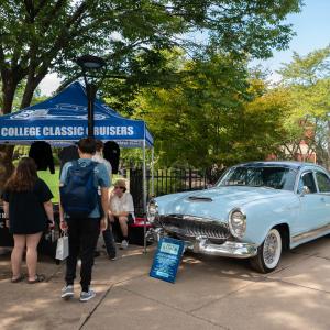 Students gathera round a table under a canopy that says "Penn College Classic Cruisers." Next to the canopy is a shiny light blue antique car with chrome accents.