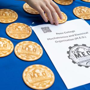 Several wood coasters that are laser-etched with the Penn College Mechatronics and Electrical Organization’s logo sit on a table with a blue tablecloth. A piece of paper on the table shows the name of the organization.
