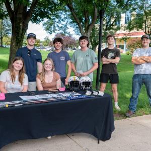 Five students stand and two sit behind a table that has hockey sticks and helmets, can cozies that say "Penn College Hockey Club" and sheets of information.