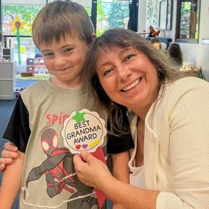 A woman poses with a preschool-aged child. She is holding a medal crafted by the child that says "Best Grandma Award."