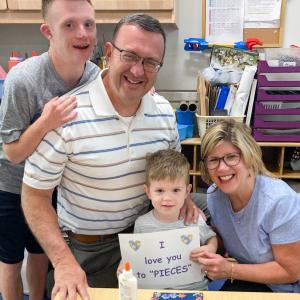 A preschool-aged child poses with family members. He holds up a sign that says "I love you to pieces." On the table in front of them is a craft made with blue paper and puzzle pieces.