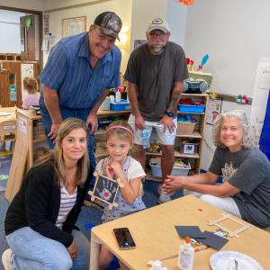 Four adults pose with a little girl who is sitting at a table and holding up a craft that shows her handprint.