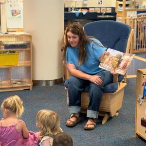 A woman sits in a rocking chair in a preschool classroom and reads a picture book to children sitting on a carpet in front of her.
