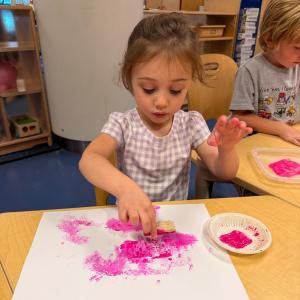A preschool-aged child uses a sponge brush to add bright pink paint to a sheet of white paper. Another child doing the same thing is visible in the background.