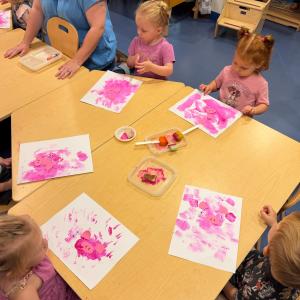 Toddlers sit in chairs at a table. In front of each child is his or her artwork: pink paint on white paper with googly eyes and pink paper pig snouts and ears.