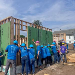 Nine volunteers hold an exterior wall up as it is braced. Other volunteers are visible holding up other walls and doing other tasks.