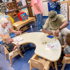 Two children sit at a table. Each works with an adult to glue pieces of construction paper onto a pieces of paper that is printed wtih a heart and the words "You have a big piece of my heart. Happy Grandparents Day!"