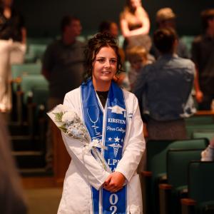 A student wearing a white lab coat and blue graduation stole with her first name and various symbols of the nursing profession walks down an aisle between auditorium seats.