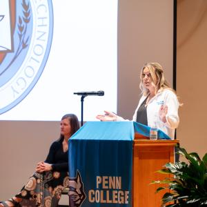 A woman wearing a white lab coat stands behind a lectern on a stage.