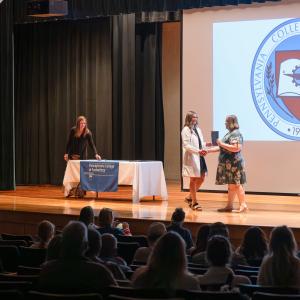 A graduate wearing a white lab coat receives a handshake and diploma folder on stage.