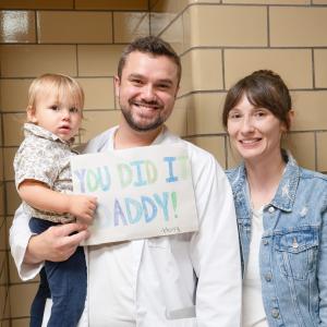 A man in a white lab coat holds a small boy and a handmade sign that says "You Did It, Daddy!" A woman stands next to them.