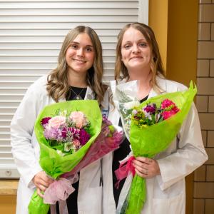 Two women in white lab coats hold bright bouquets.