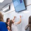 Two women write on a white board, with others gathered around them.