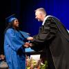 A student in graduation regalia shakes the hand of President Reed, also in a graduation gown, on the Community Arts Center stage.