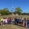 Children line up on a lawn with a large American flag hanging on a pole in the background.