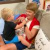 A toddler looks at his grandmother while holding her hands and sitting on her lap on a couch.