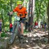 A boy rides a bike down a rocky, wooded path while onlookers stand along the edges of the trail.