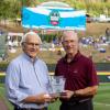 State Sen. Gene Yaw and Little League International Board of Directors Chairman Steven P. Johnson stand at the edge of a baseball field holding a glass trophy.