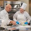 In Penn College's baking lab, a man in a chef's uniform provides instruction to a young woman, also in a chef's uniform, as both look at a product on a stainless steel table. Commercial ovens are behind them.
