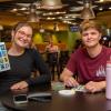 Two students sit at a table in Penn College's CC Commons. One is holding up a loteria board. The other has a board sitting in front of him. A cup of bingo markers sits between them to mark their boards.