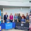 Penn College President Michael J. Reed sits behind a table with Penn College signage, and Thaddeus Stevens College of Technology President Pedro A. Rivera II sits behind a table with Thaddeus Stevens signage. A group of people from both colleges stands behind them.
