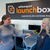 Erin Shultz speaks into a microphone held by WNEP reporter Mackenzie Aucker. They stand in front of a large wall sign that reads Williamsport LaunchBox supported by Ken and Kristie Healy.