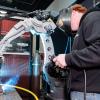 A Pennsylvania College of Technology student operates a robotic welding system. The student is wearing a welding helmet. Light and smoke emanate from the end of the robotic arm, which is touching metal.