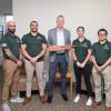 Penn College President Mike Reed stands in the center of a group of students holding a wooden sword. The students are all wearing green shirts and tan pants.