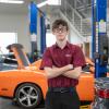 Uniformed automotive student stand with arms crossed in lab in front of orange mustang.
