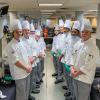 Students in chefs uniforms stand along a buffet.