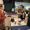 Three people stand at a table with equipment in an indoor exhibit space.