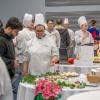 People gather around tables in Penn College's Thompson Professional Development Center. Near the center of the photo, a student in a chefs uniform gestures toward pastries on her table while talking with a young man.
