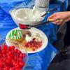 Close-up of a plate with donuts, whipped cream, strawberries, and ice cream, while a hand adds cherries from a spoon; bowls of cherries and whipped cream are visible on a blue-covered table.