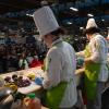 Students in chefs uniforms with green aprons prepare food on the Pennsylvania Farm Show’s Culinary Connection stage. Various vegetables are arranged on the counter. A seated audience watches the cooking demonstration in a large indoor venue with booths and banners in the background.