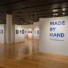 Artwork hands on walls in The Gallery at Penn College. One wall shows exhibit name, "Made by Hand," in large blue letters.