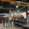 Four people standing in Penn College’s Lycoming Engines Metal Trades Center beneath an overhead crane, surrounded by welding equipment, fabrication tools, and a plasma cutting machine.