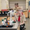 A boy in a Scouting uniform and safety goggles sits at a bench in a chemistry lab.