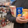 Close-up of an arm holding a box of instant mashed potatoes, a box of macaroni and cheese and a jar of peanut butter. Shelves lined with food are visible in the background.