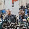 Three people stand near a large diesel engine, one looking at a gauge.
