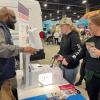 An adult demonstrates a laparoscopic surgery simulator to two participants at a booth, where visitors test controls and observe a lighted display inside a busy expo hall.