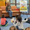 A woman sits on a couch with a book in her hand while children sit on the floor looking at her.
