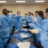 A recent alumna points while demonstrating a room full of students how to properly put on gowns and gloves.