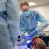 A dental hygiene student in protective medical gear shines a blue light into a patient’s mouth while another student observes.