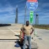 Dwight Alexandar stands under a sign that says 'Road to Space.' Behind him is a rocket launchpad.