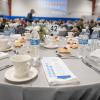 A table set with a tablecloth, plates, cups and a cloth napkin. On top of the napkin is a Scholraship Luncheon program. In the background, the Penn College Field House is filled with similar tables surrounded by guests.