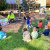 A man in a flourescent hoodie points to pictures of nature objects on a poster board as toddlers sit in the grass watching.