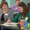 Adults sit at a table in a conference room taking notes.