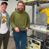 Two students stand next to a Fanuc robot, one holding the robot's controller. They are in Penn College's Process Control and Robotics Lab.